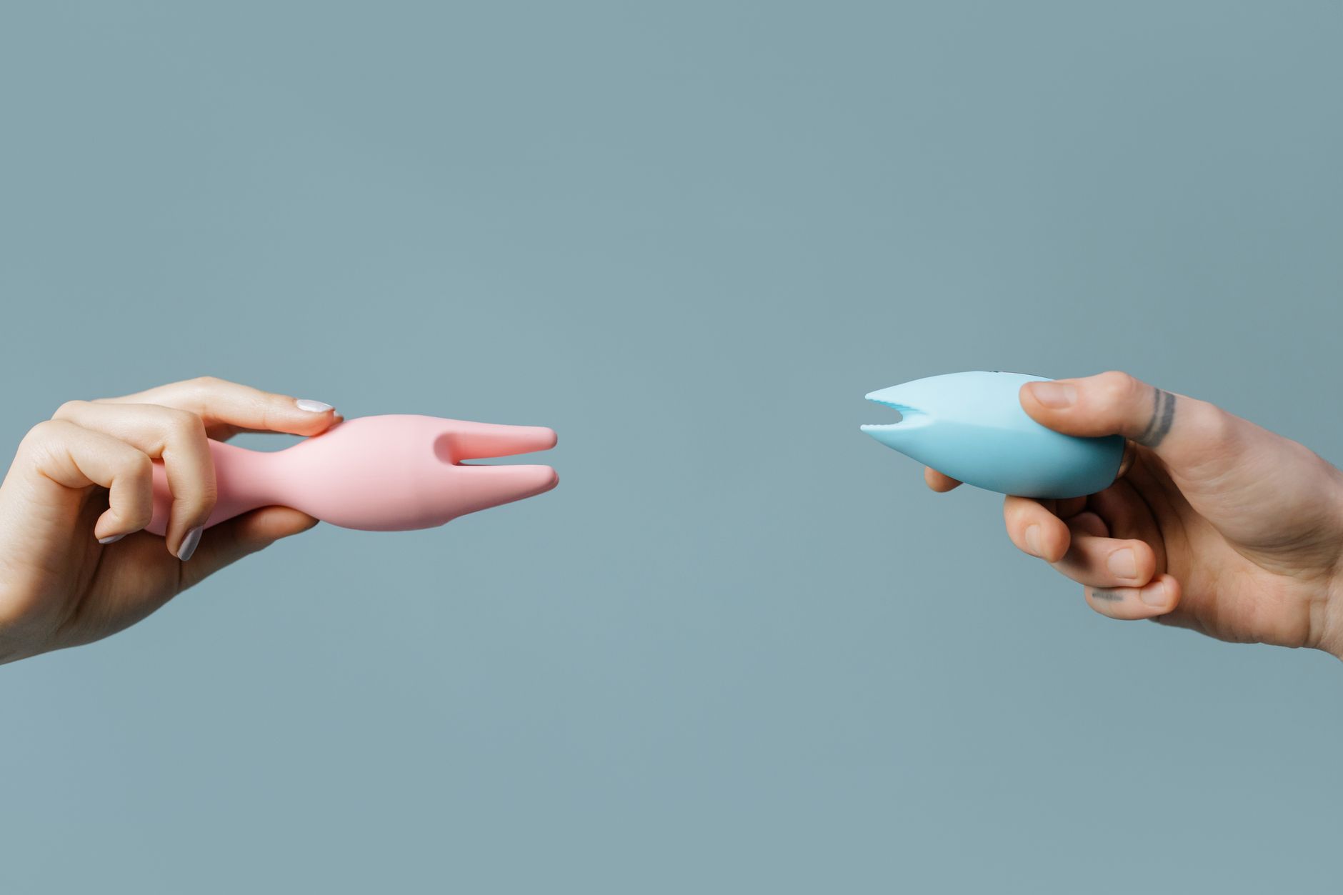 Deux mains tenant des vibromasseurs rose et bleu contre un fond pastel minimaliste